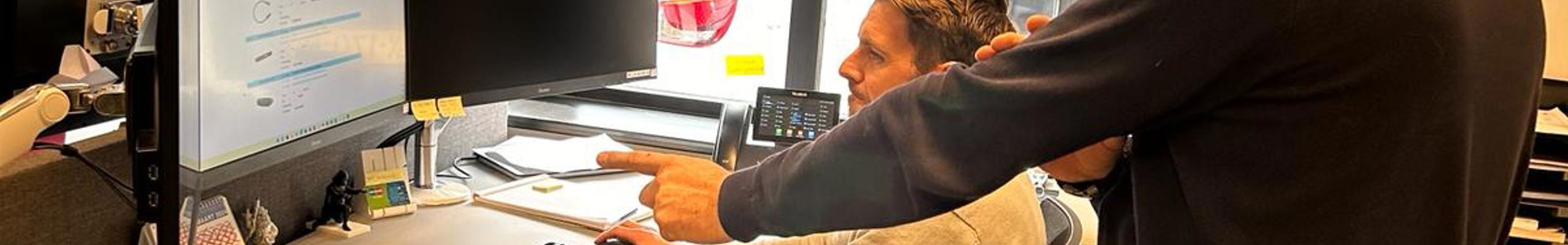man geeft staand aanwijzingen aan andere man zitten voor een beeldscherm