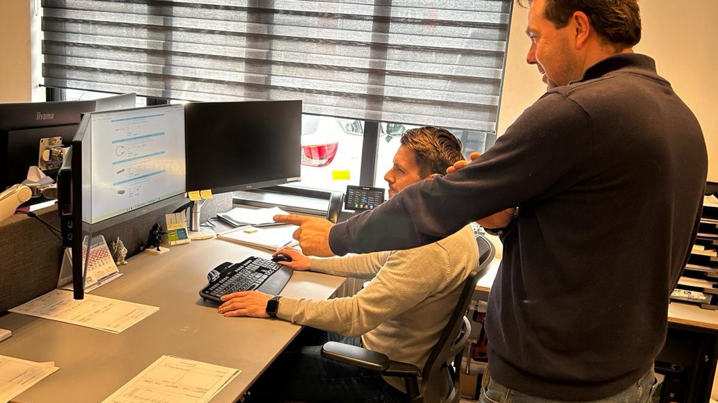 man geeft staand aanwijzingen aan andere man zitten voor een beeldscherm
