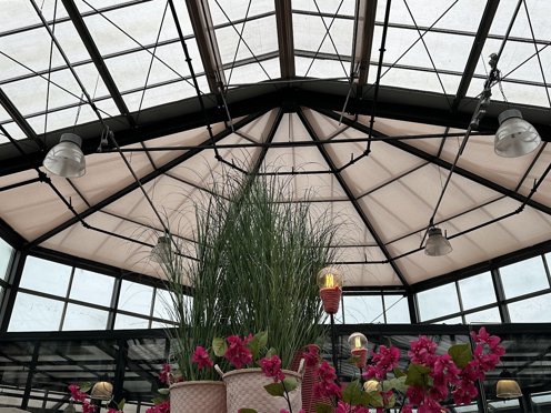 schermdoek in de nok van een ronde glazen hal van een tuincentrum met roze planten op de voorgrond
