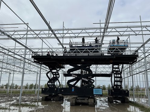 drie hoogwerkers in de modder met mannen in de hoogte aan het werk aan een kas in aanbouw