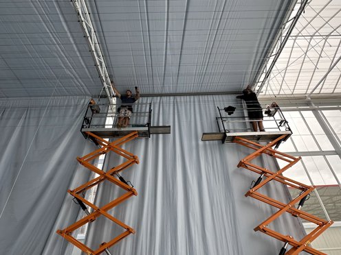 twee oranje hoogwerkers helemaal op zijn hoogst met twee mannen die schermdoeken installeren in een tuinbouwkas